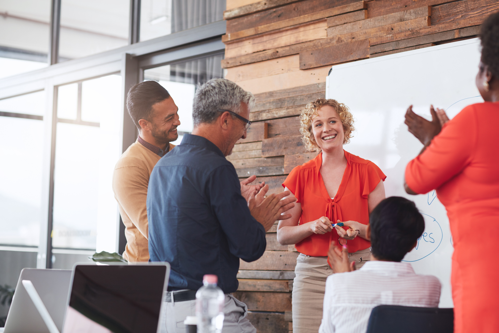 group of five co-workers stood or sat around a white board clapping