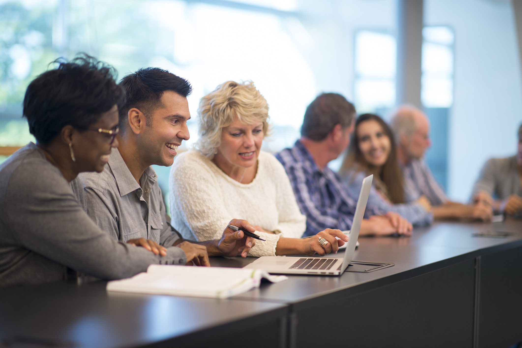 a diverse range of people, of all races and ages, sat together working and looking at a laptop